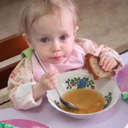 Young girl holding a piece of bread and eating soup out of a bowl with a very large spoon