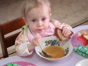 Young girl holding a piece of bread and eating soup out of a bowl with a very large spoon
