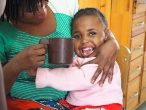 Young girl hugging her caregiver and smiling while her caregiver holds a mug