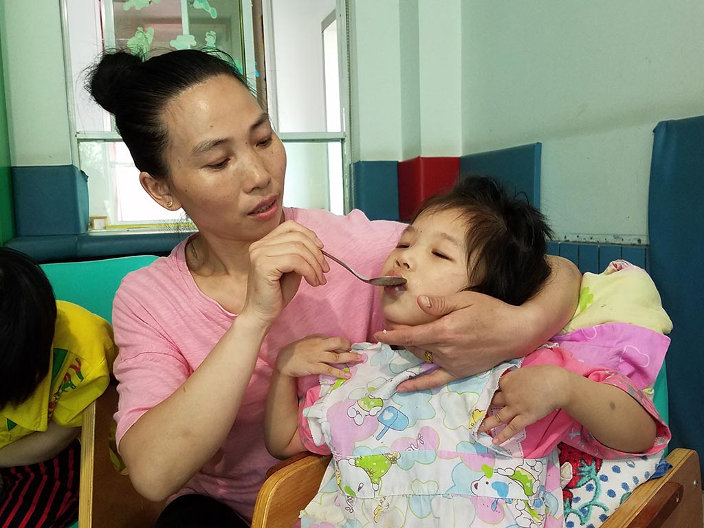 Caregiver feeding a child in a high chair with a spoon and supporting her face