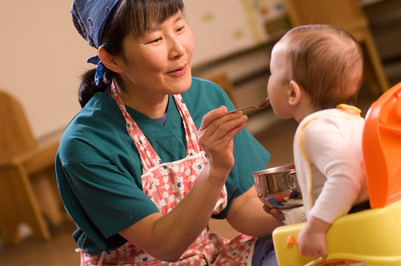 Caregiver feeding an infant who is sitting in a high chair using a spoon