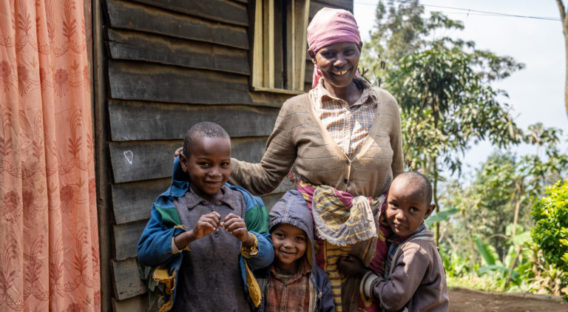 Woman in Tanzania standing outside of a wooden house with her three smiling sons