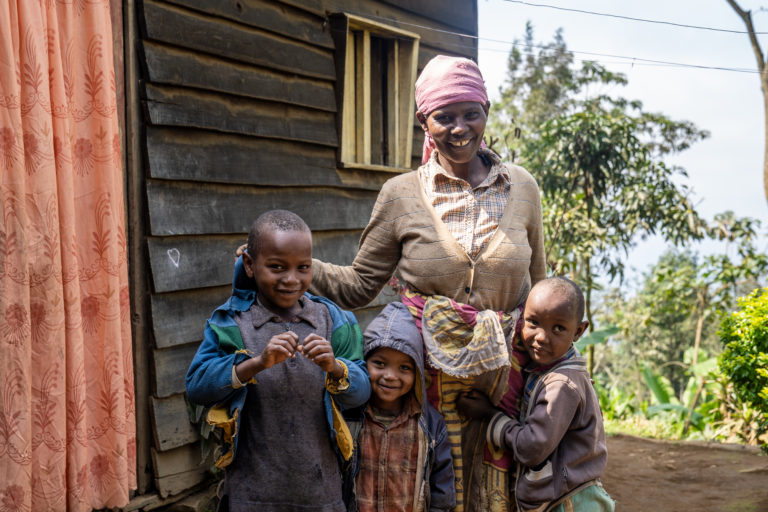 Woman in Tanzania standing outside of a wooden house with her three smiling sons