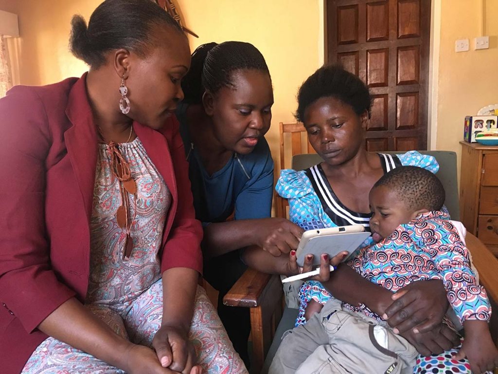 Two women showing a mother holding her child on her lap something on a tablet