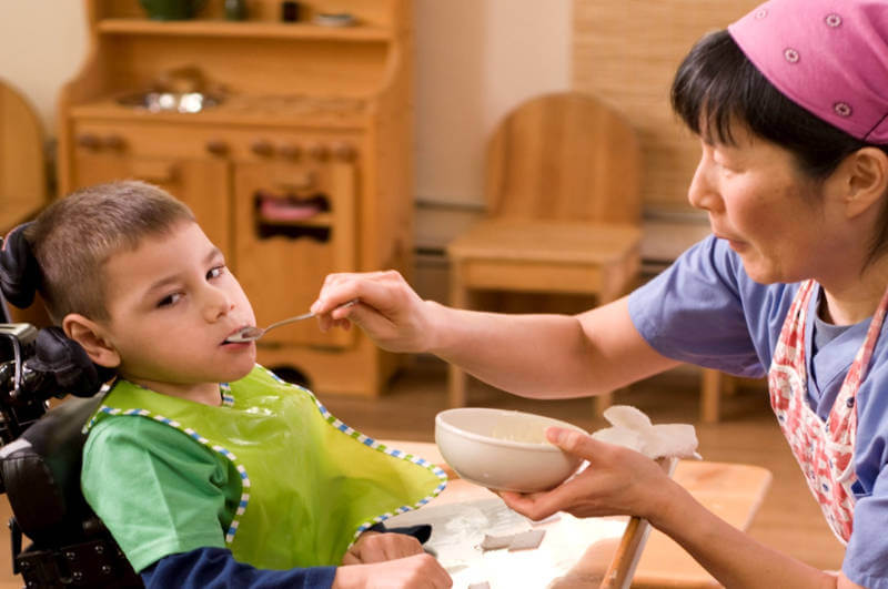 A caregiver feeding a young boy with disabilities who is in a wheelchair using a spoon
