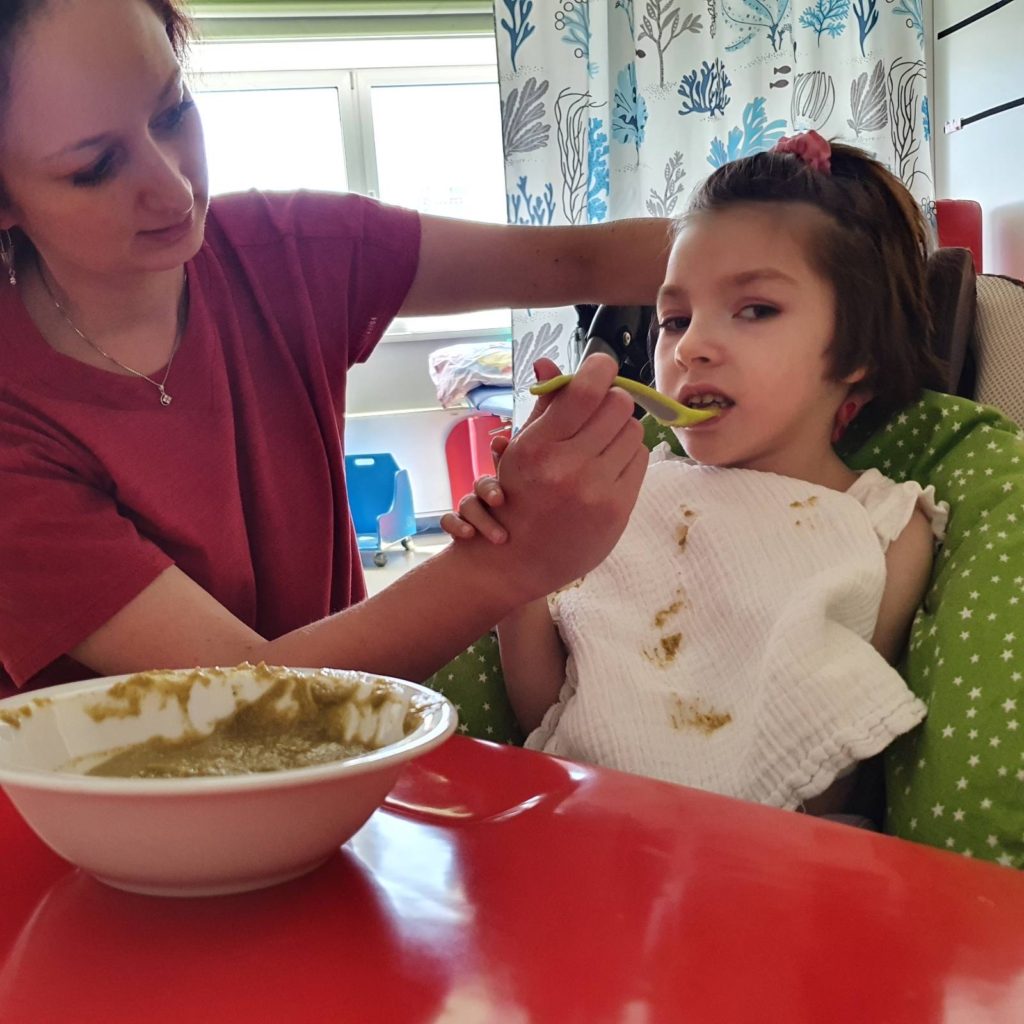 Young girl with a disability in a high chair being fed with a spoon by a caregiver