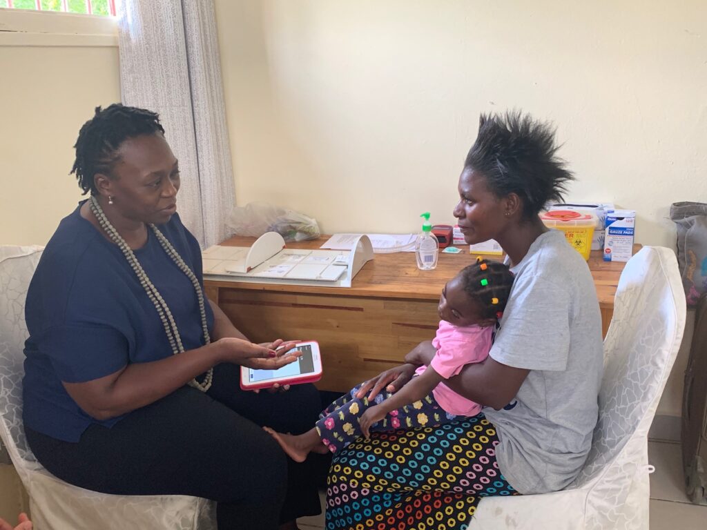 A Count Me In user speaking with a mother of a child with disabilities while holding a phone showing with the app up. The child is on her mother's lap. They are Zambian.