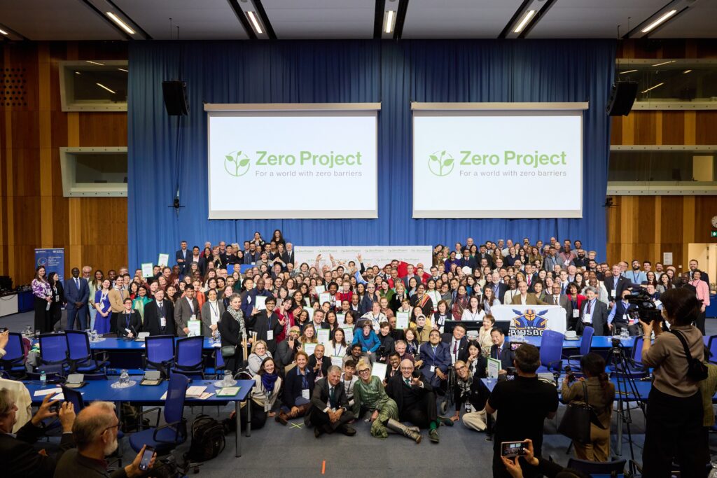 A group photo of attendees of the Zero Project conference in 2024 in front of screens advertising the conference