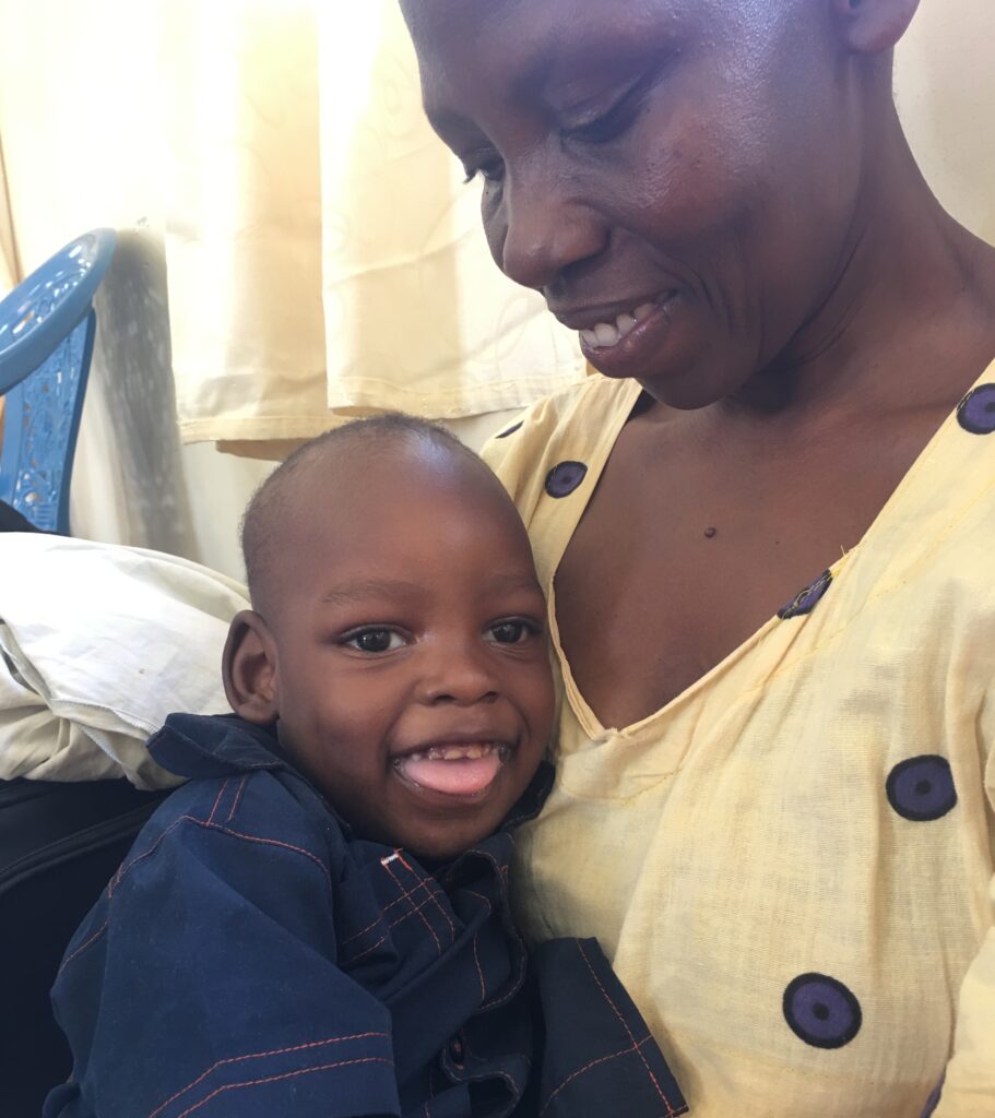 A young child with disabilities in Uganda sits on his mother's lap and both are smiling.