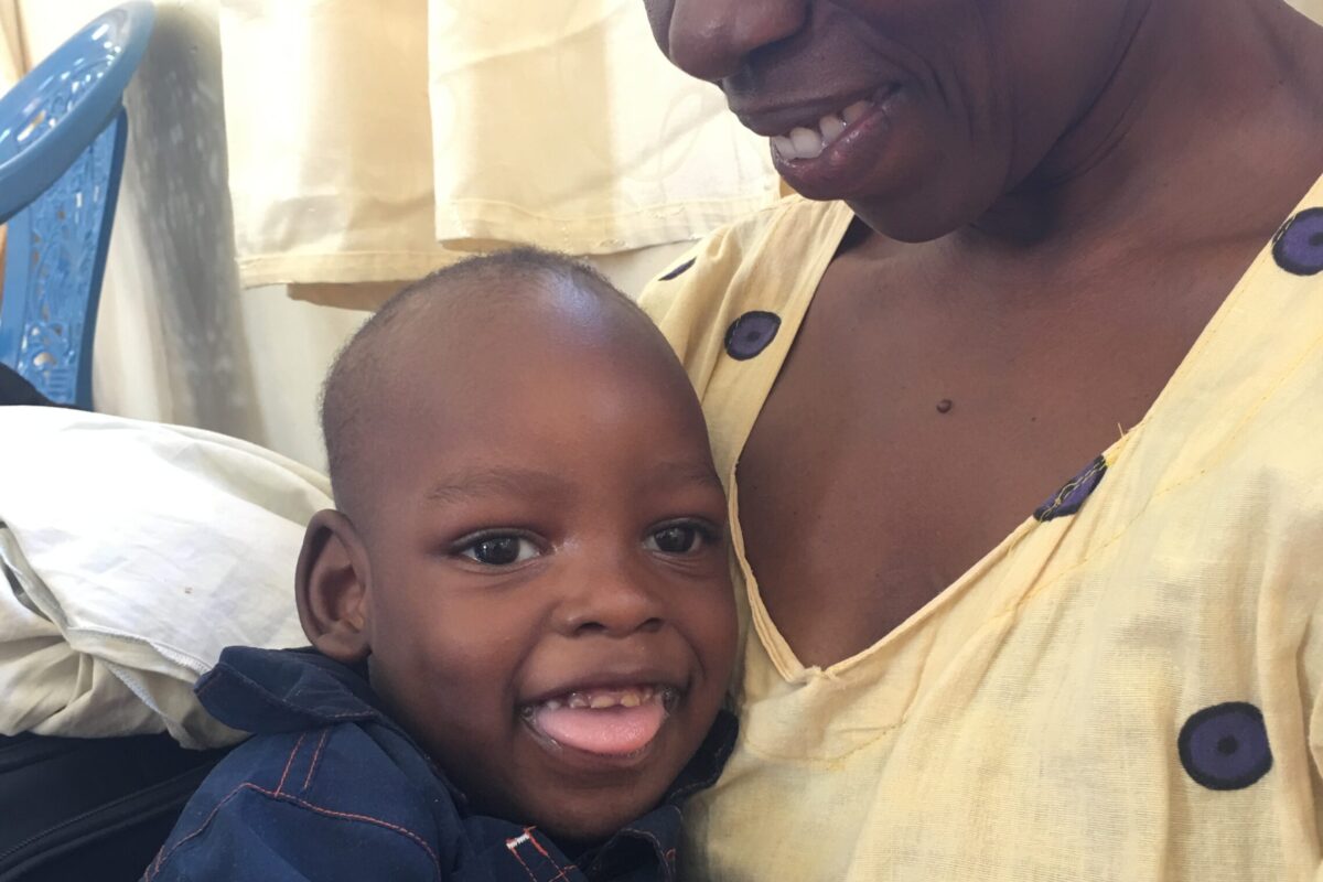 A young child with disabilities in Uganda sits on his mother's lap and both are smiling.