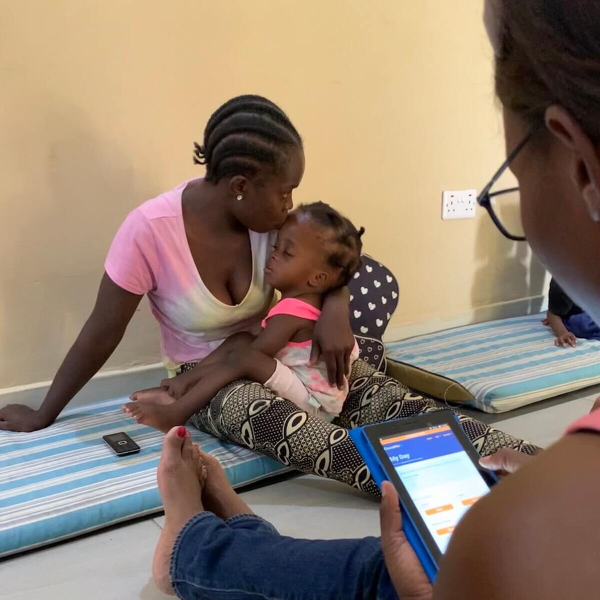 A mother holds her child while a person looks on with SPOON's app Count Me In pulled up on a phone.